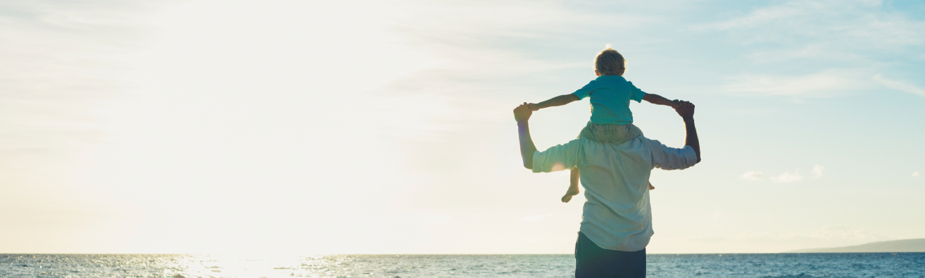 A boy on his father's shoulders looking out at the ocean on a beach.